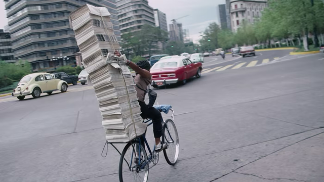 Bicyclist in traffic, right arm reaching behind and up to steady a 7-foot stack of newspapers banded to the rear rack