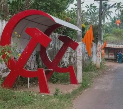A bus stop with two giant red hammer and sickle symbols on the side holding up the roof of the bus stop with a bench underneath.