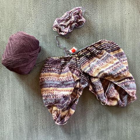 Two knit triangles connected to each other on a large circular needle. They're shown on a speckled grey background, together with a ball of variegated yarn in various lilacs and a ball of aubergine colored yarn.