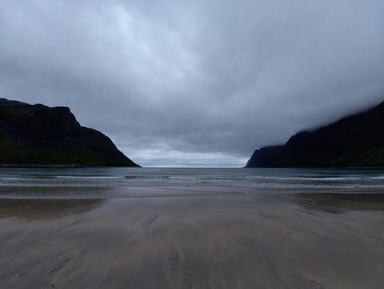 Ein Strand bei Ebbe, rechts und links hohe Berge, die teilweise in den tiefhängenden Wolken verschwinden.