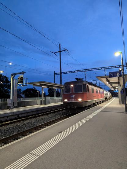 Une locomotive ancienne 620 de CFF Cargo traverse la gare de Rolle en tête d'un train de marchandises composé de wagons citerne. Le ciel est bleu nuit.