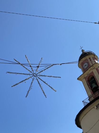 An einem Kabel über der Straße ein einfacher, großer Stern mit 10 Streben und Lämpchen. Rechts daneben ein klassizistischer Kirchturm, blauer Himmel.