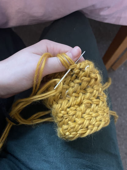 Hand, with yarn wrapped around the thumb and a needle pushed through the loops holding a piece of textile