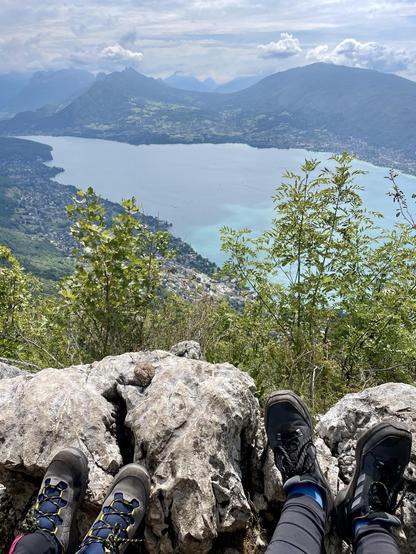 A scenic view from a rocky perch overlooking a lake surrounded by mountains, with two pairs of hiker's feet in foreground.