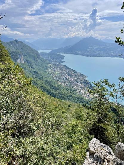 A scenic view of a mountainous landscape overlooking a lake, with a small town nestled along the shoreline, surrounded by lush greenery and distant mountains under a partly cloudy sky.