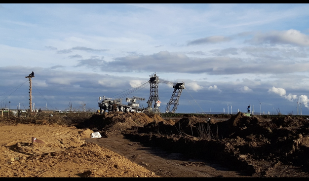 Ein Foto direkt in der Nähe. Das riesige baggers. Man sieht zwei seiner Arme. Der Bagger steht still. Links  ein holzpfahl, auf dem ein Mensch sitzt, ein monopod.
