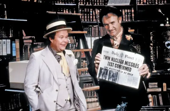 Publicity still from the 1978 film "Superman: The Movie," picturing villain Lex Luthor holding a newspaper with a headline about a live nuclear missile test, as his stooge Otis (Ned Beatty) smiles and looks on appreciatively.