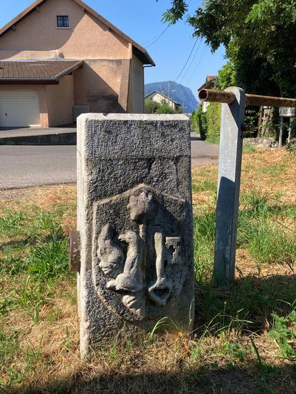 A Swiss / French border stone showing the Geneva shield of an eagle and key engraved next to a rusting barrier. A house is in the background.