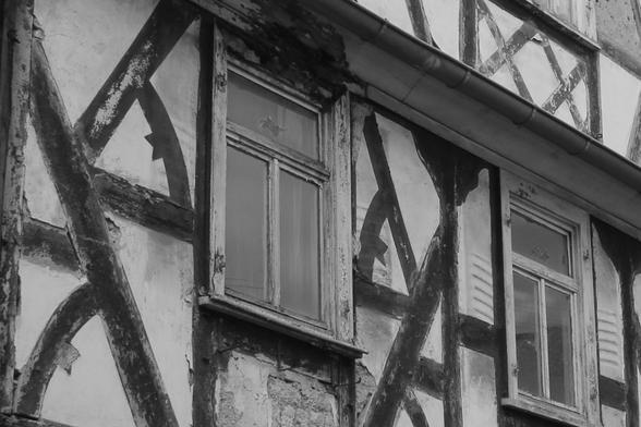 Bild in schwarz-weiß: Fassade eines Fachwerkhauses. 
Die Farbe der Gefache, Balken und Fensterrahmen blättert. Manche Gefache liegen frei. Die Fensterläden fehlen. Das Gebäude steht leer und ist in einem schlechten Zustand.
