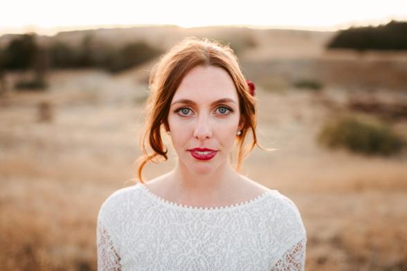 This image features a close-up portrait of a woman standing outdoors in a natural, dry landscape. The background appears to be an open field with soft, golden light, likely taken during the golden hour near sunset. The woman has light skin, blue-green eyes, and medium-length, reddish-brown hair styled in a loose updo with a few strands softly framing her face. She is wearing a white lace dress that has a delicate, intricate pattern and a round neckline adorned with tiny pearls. Her expression is neutral, yet calm and serene, with her gaze directed straight at the camera. The overall mood of the image is warm, peaceful, and intimate, highlighting the subject against a softly blurred, natural backdrop.