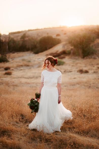 This image shows a full-body portrait of a woman in a bridal gown standing in a dry, open field during sunset. The landscape is golden and serene, with rolling hills in the background and soft sunlight illuminating the scene.

The woman is wearing a white, lace wedding dress with a fitted bodice and short sleeves, paired with a flowing tulle skirt. Her hair is styled in a loose, elegant updo with a few tendrils softly framing her face, and she has a red flower tucked into her hair on one side. She holds a bouquet of flowers in one hand, but instead of holding it up, she carries it down by her side, allowing the flowers to naturally complement the scene.

She gazes downward, with a soft and contemplative expression, while a gentle breeze seems to catch her hair and dress. The overall mood of the image is peaceful and introspective, with the natural, warm tones of the setting sun enhancing the tranquil atmosphere. The scene captures a moment of quiet beauty, with the bride standing gracefully amidst the vast, rustic landscape.