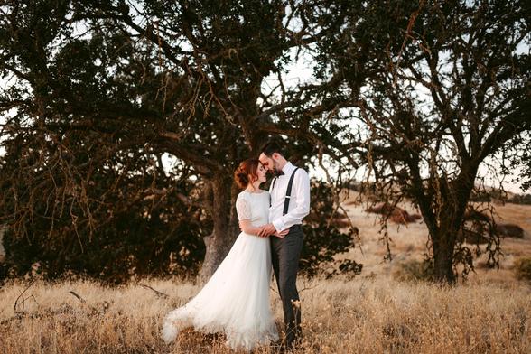 The image depicts a couple standing closely together in a dry, grassy field under the shade of a large, gnarled tree. The woman, dressed in a white lace wedding gown, leans into the man, who is wearing a white shirt, suspenders, and gray trousers. They are embracing tenderly, with the man's forehead resting gently on the woman's, creating an intimate and peaceful moment. The scene is set in a natural, somewhat rustic landscape, with the warm tones of the dried grass and the soft evening light adding to the romantic and timeless atmosphere. The background is softly out of focus, drawing attention to the couple as the central element of the image.
