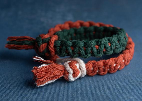 A photograph of a pair of cordage bracelets tied up with Portuguese sennit in 3mm cotton cord, one rust on white, the other green on rust. 
