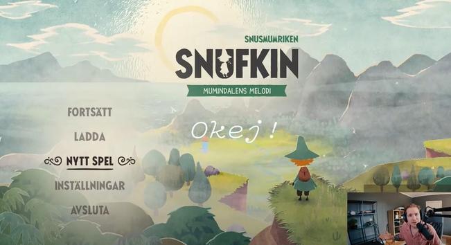 Screenshot of a YouTube video moment, showing the title screen of the game "Snufkin - Mumindalens Melodi" [Snufkin - Moomin valley's melody]. In addition to some menu options (Fortsätt, Ladda...) we see Snufkin standing on a cliff with his back turned to us, looking down at the valley with the moomin family's house in the distance, the sea and grey mountains to the left, and grey steep mountains enclosing the green and lush valley from the right. The sun is rising (I assume) bright and yellow in the centre and is fully seen above the horizon.
At the bottom right we see a video cam picture with a white male, sitting at a desk, light red / dark salmon shirt, dark blond hair, looking at the camera/ us with a friendly inviting face.