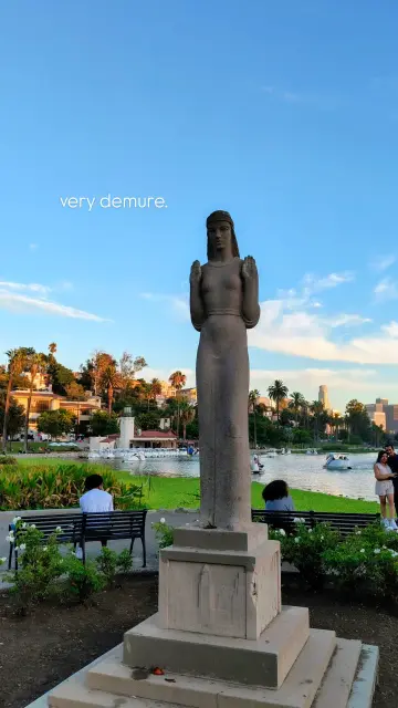 The 'Lady of the Lake' statue at Echo Park Lake, with swan-shaped pedal boats in the water behind it and the late-afternoon sun highlighting various buildings in the distance.