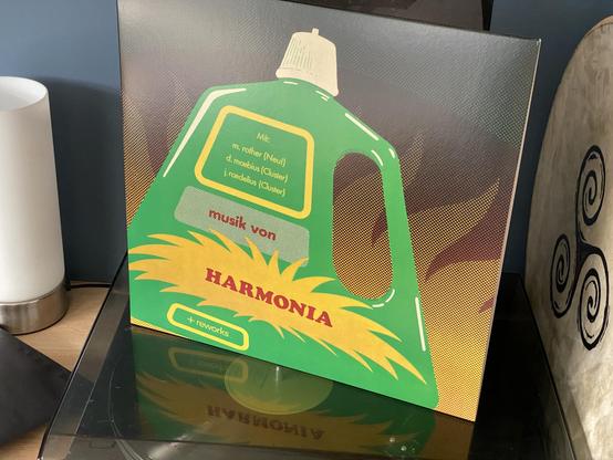 The image shows a vinyl record cover featuring a graphic design of a green bottle with yellow accents. The text includes the title "HARMONIA," names associated with the artists, and the phrase "musik von." The cover is placed on a surface