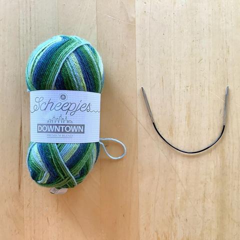 A ball of self striping sock yarn in green colors is shown together with a tiny circular knitting needle on a light wooden background.