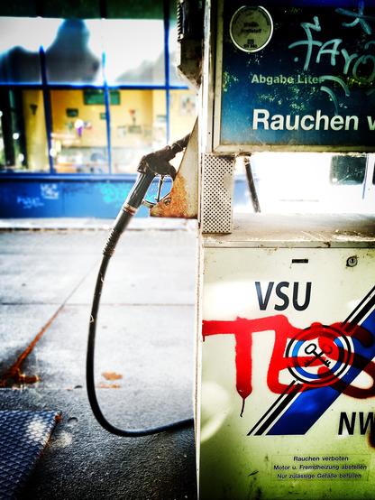 Foto zeigt eine stillgelegte Tankstelle an einem sonnigen Tag, Fensterfront in blau kontrastiert mit dem orangegelb gestrichenen Innenraum. Innen an der Wand hängen alte Bilder und Kalender. Die Zapfsäule ist verrostet, mit Graffiti besprüht, und dreckig.