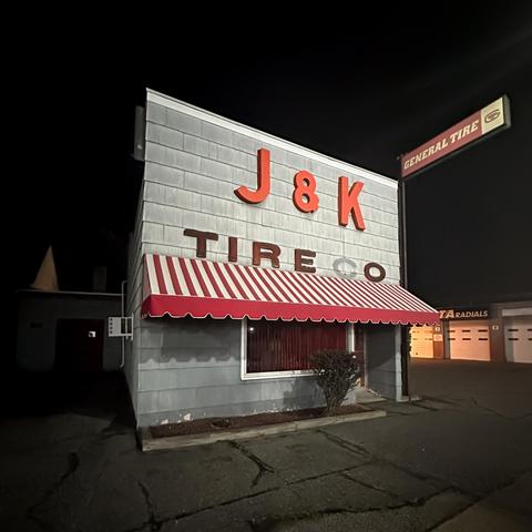 Color photo of a small isolated building with a square frontage to the street. It is photographed at night from a slight angle so that the facade takes on a more trapezoidal shape. It is illumined by a street light that is out of frame. The name of the business is displayed above a red and white striped awning in two lines in capital letters, the upper set larger than the lower. “J & K” resides above “TIRE CO” - the “c” in “CO” is missing but the shingles around the missing letter have faded more than where the letter protected them from the sun. 