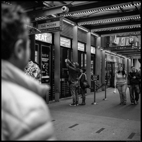 A couple stands in front of a theater ticket window beneath string lights and signs.  One is raising a phone in both hands to take a picture.  A blurry person walks towards them in the foreground while two others are set to pass by in the background.