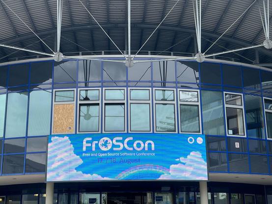 Foto vom Eingang eines Konferenzgebäudes, Glasfront. Über der Tür ein Display mit dem Logo der FROSSCon,  darunter der Schriftzug "Free and Open Source Software Conference 17./18. August"