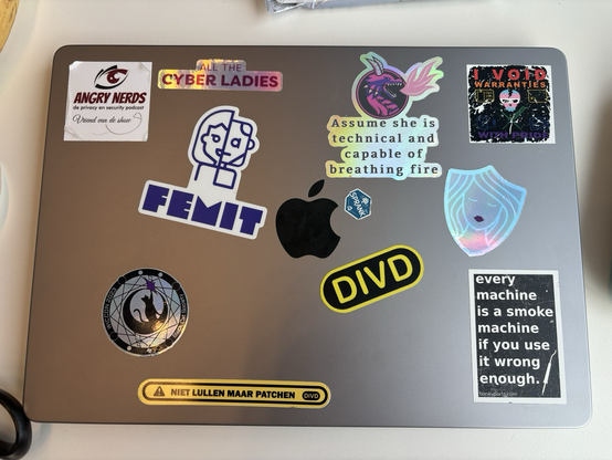 MacBook with stickers: a stray sticker from an actual apple, “assume she is technical and capable of breathing fire” by MiFare_lady, “all the cyber ladies” by Lucinda Sterk, “I void warranties with pride” by Jilles, Wiccon 2023, Angry Nerds Podcast, DIVD, “niet lullen maar patchen” and “every machine is a smoke machine if you use it wrong enough” from blinkyparts