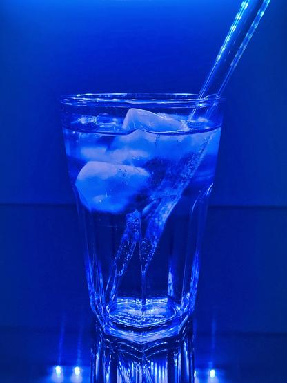 Ein Longdrink-Glas mit Gin Tonic, Eiswürfeln und Glasstrohhalm steht auf einer Glasplatte und wird von blauen LEDs sphärisch beleuchtet. 