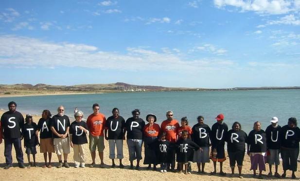 People wear t-shirts with letters spelling out

STAND UP FOR THE BURRUP 

On sand at beach in Pilbara region