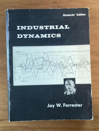 Black book on a wooden table:  Upper corner small letters “Student’s Edition”,  Left justified title

INDUSTRIAL
DYNAMICS

Then a white plot of undulating curves solid, dashes and such,  a small photograph of a man and below that

Jay W. Forrester