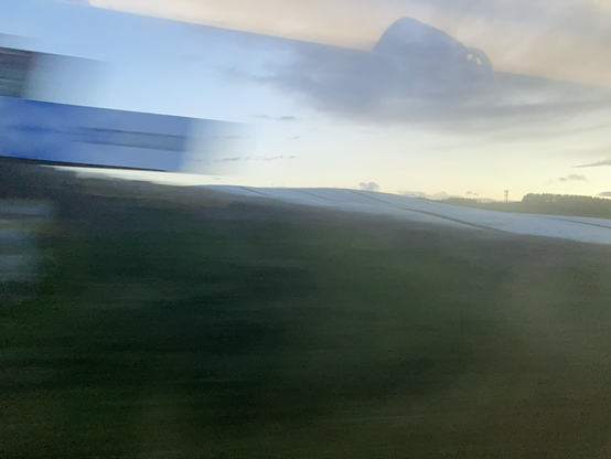 Very speed blurred photo with the Scotland “arm” of the border sign alongside the East Coast railway line only just visible moving out of shot, in front of a similarly blurry grassy field 