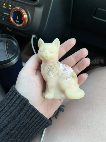A figurine of a sitting cat made of a creamy rose and yellow glass, painted with flowers on its back