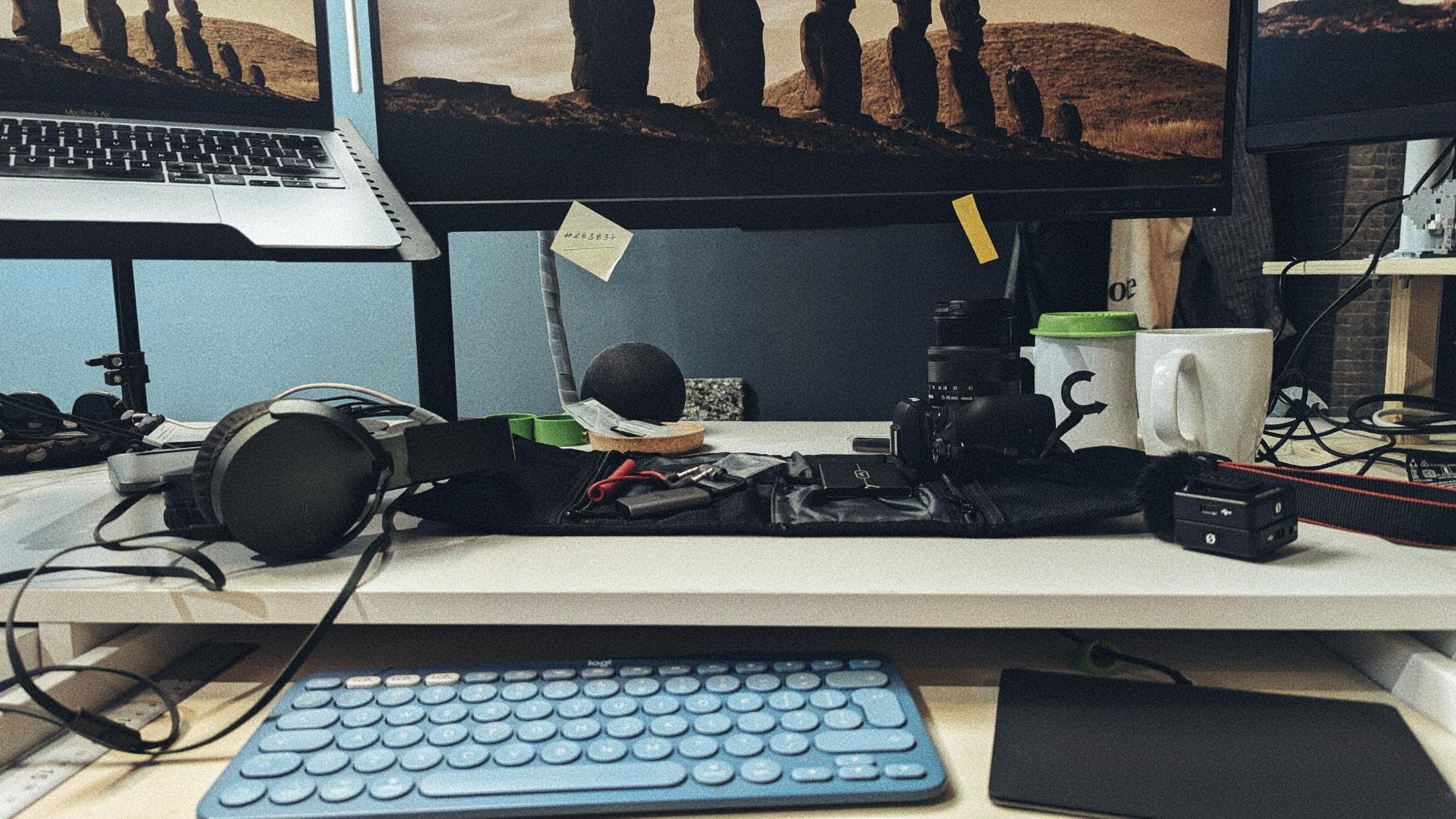 A cluttered workspace features a keyboard, headphones, camera, lens, mug, notepad, and multiple computer monitors.