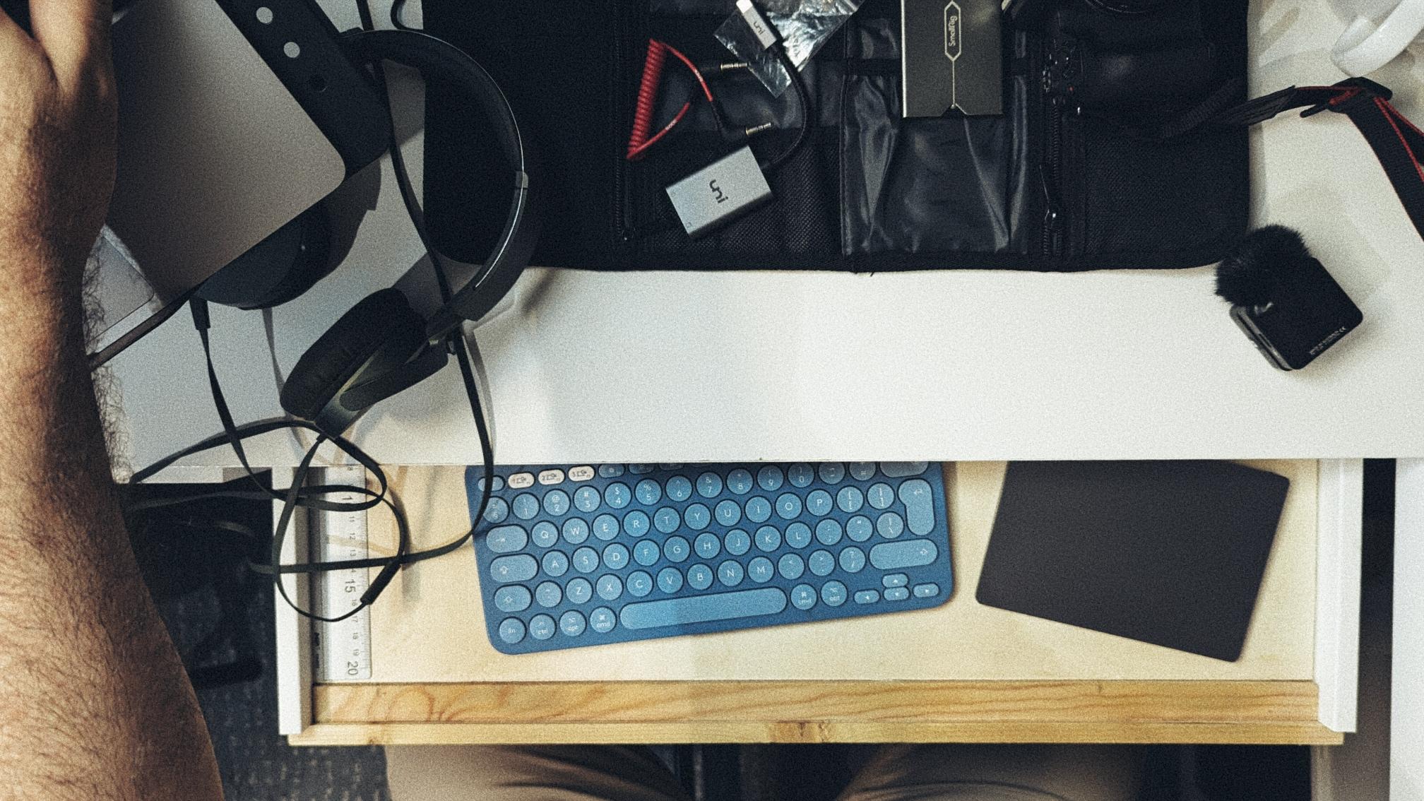 A person is sitting at a cluttered desk with various electronic devices, including a keyboard, headphones, and other tech accessories, in view.