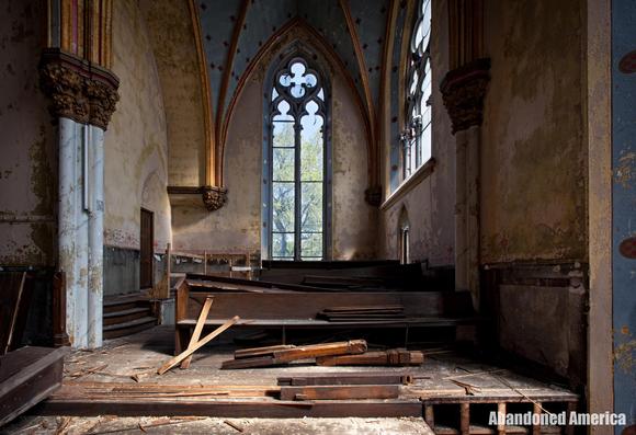 A battered pew in the choir loft of a church. Boards are littered on the floor. Behind the pew is a tall, narrow window and a tree is visible outside. 