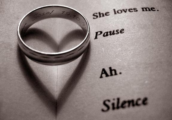 S/W-Bild eines Eherings mit Gravur „Josef 19.10.79“ , der mittig in einem aufgeklappten Theaterskript liegt. Der Schatten des Rings formt ein Herz, auf der rechten Seite des Skripts steht geschrieben „She loves me. Pause. Ah. Silence.“