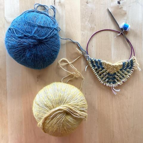 The start of a triangle shawl on a long circular needle, shown together with a ball of yellow and a ball of petrol colored yarn on a light wooden background.
