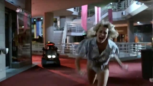 A woman with disheveled hair is running in a corridor, appearing frightened. Behind her, a small vehicle with lights is pursuing her. The setting features red carpets, staircases, and colorful decorations.
