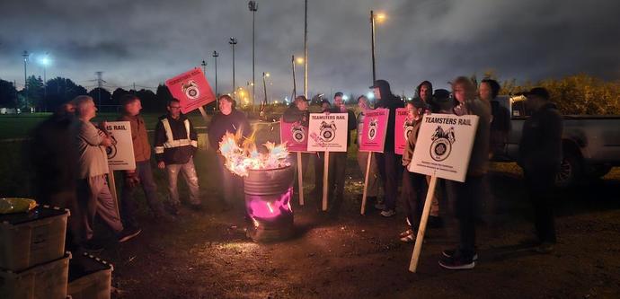 
Canadian rail workers Teamsters members were locked out because they refused to buckle to company demands for unsafe and inhumane scheduling. 

Photo: Teamster union rail workers gathering around a fire in a drum under night sky