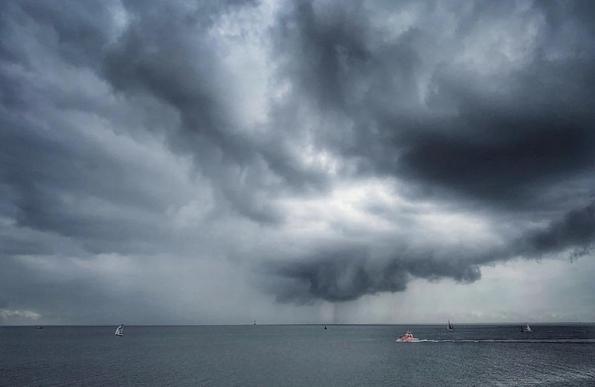 Dramatischer Himmel mit dunklen Wolken und Gewitterzelle samt Starkregen an der Nordsee. Vom rechten Bildschirmrand fährt ein kleines rot-weißes Schiff der Seenotretter ins Bild. 