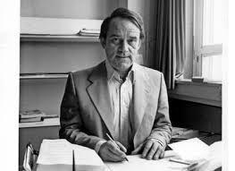 Helmut Kentler slightly grinning as he sits at his desk, pen poised over a notebook as if he was caught writing.