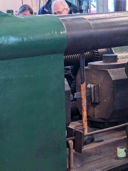 Looking at the sample being tested in the machine. Between massive steel castings you can just see a fractured metal bar. The shaft of a hammer leans up against it. 