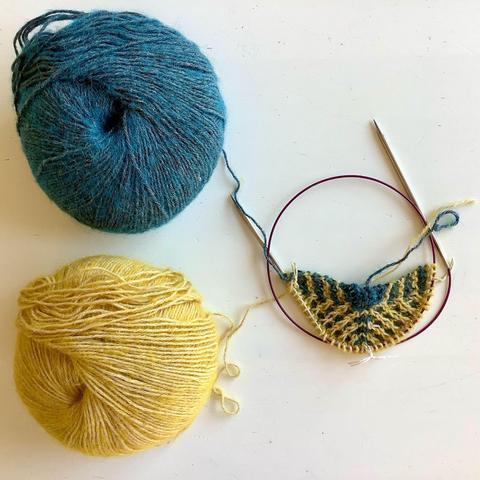 The start of a triangle shawl in mosaic patterning on a circular needle. It is shown together with a ball,of yellow and a ball of petrol colored yarn on a white background.