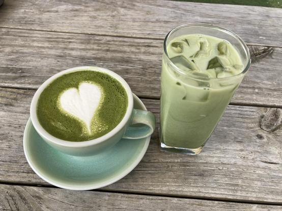 Two cups of matcha latte on a wooden table, one is iced in a tall clear glass, the second is hot with a decorative white heart drawn in to the green foam.