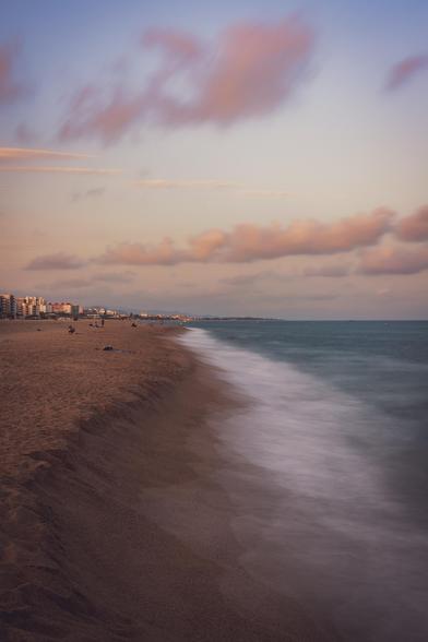 Fotografía vertical donde se ve una playa semivacía al atardecer. Mitad izquierda de la foto es la zona de tierra y la mitad derecha se puede ver el mar en larga exposición.