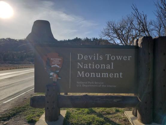 The doll ranger sarah stand in front of the sign for Devils Tower with Devils Tower in  the upper left side