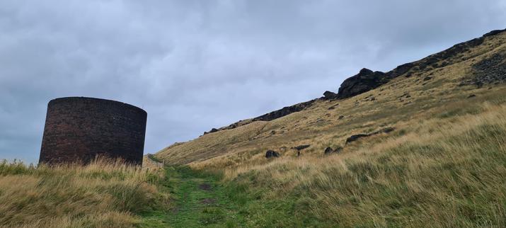 A moorland hillside, covered in grass. The grass on the slopes has gone to seed and is yellow, but a shorter green path winds through the middle. To the left of the path, there is a brick cylinder- a chimney/air vent for the canal tunnel deep under the hill. A ridge of dark rocks/cliffs lines the top of the hill.