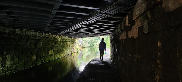 The silhouette of a tall man walking along a canal towpath, through a tunnel. The area under the bridge is dark but beyond, it's a bright day (hence the man being in silhouette). The tunnel has stone walls and its roof is made from metal girders.