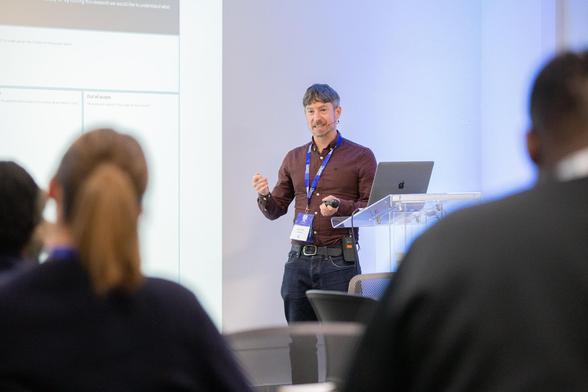 A person standing at a podium with a laptop, giving a presentation to an audience. The presenter is holding a clicker and has a name badge and lanyard.