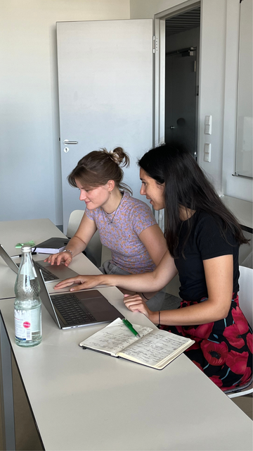 Thea und eine Kollegin sitzen am Schreibtisch. Vor ihnen stehen zwei Laptops, links und rechts liegen jeweils ein Notizbuch. Sie arbeiten gemeinsam. 