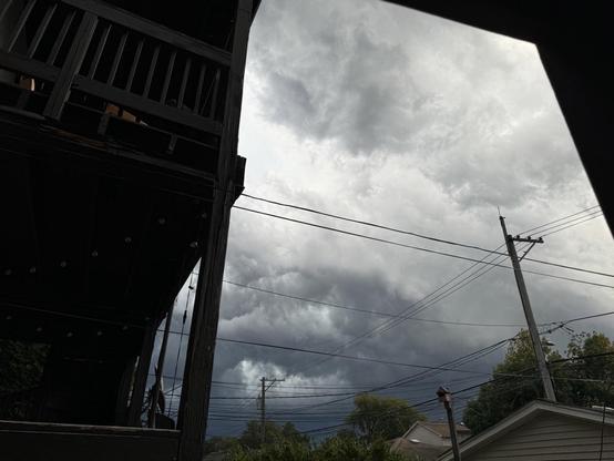 A cloudy sky is visible, featuring dark and ominous clouds. In the foreground, part of a wooden structure and utility lines are seen, along with hints of nearby buildings and greenery.